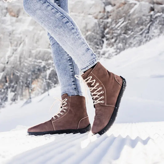 zapatos de invierno mujer Barefoot de caña alta, color marrón y con cordones blancos, caminando sobre la nieve en la montañ