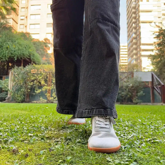 Primer plano de pies caminando en pasto con zapatillas barefoot grises.