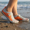 Pies de mujer en la orilla del mar usando zapatillas de agua Saguaro naranjas.