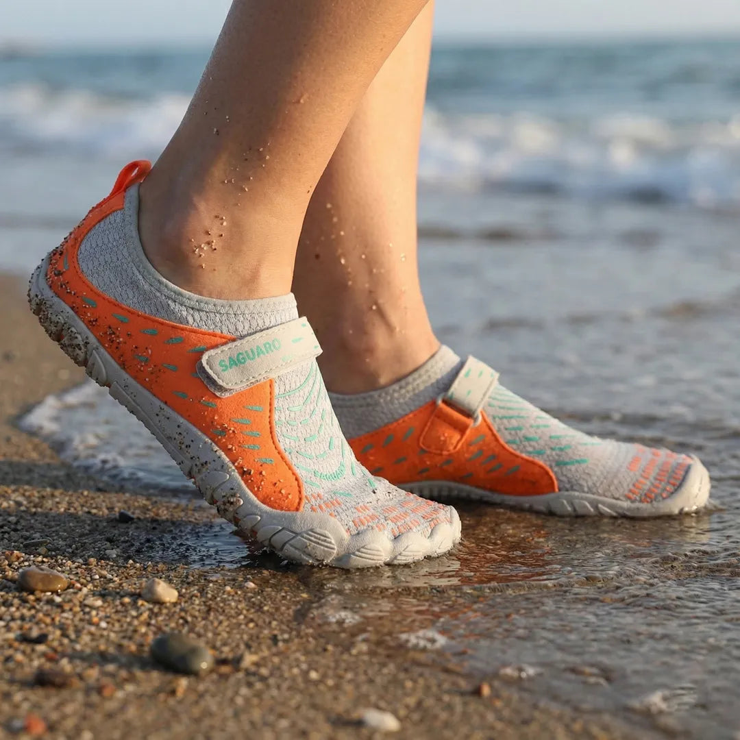 Pies de mujer en la orilla del mar usando zapatillas de agua Saguaro naranjas.