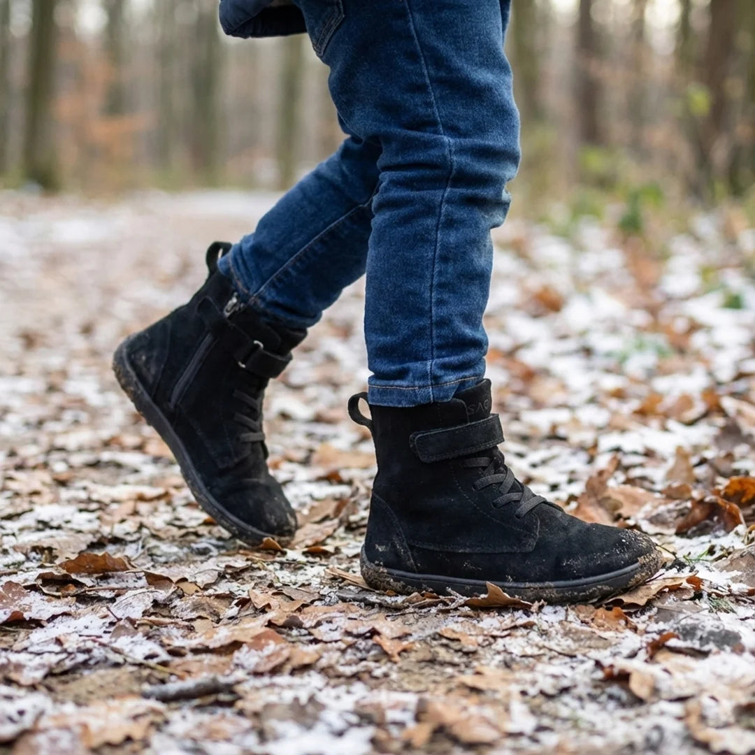 Niño caminando por sendero con hojas usando botas de invierno barefoot negras.