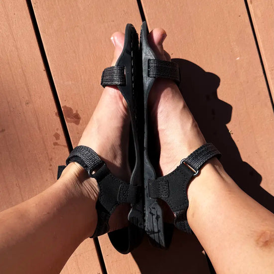 Persona usando sandalias Sowilo Raido negras sobre superficie de madera exterior.