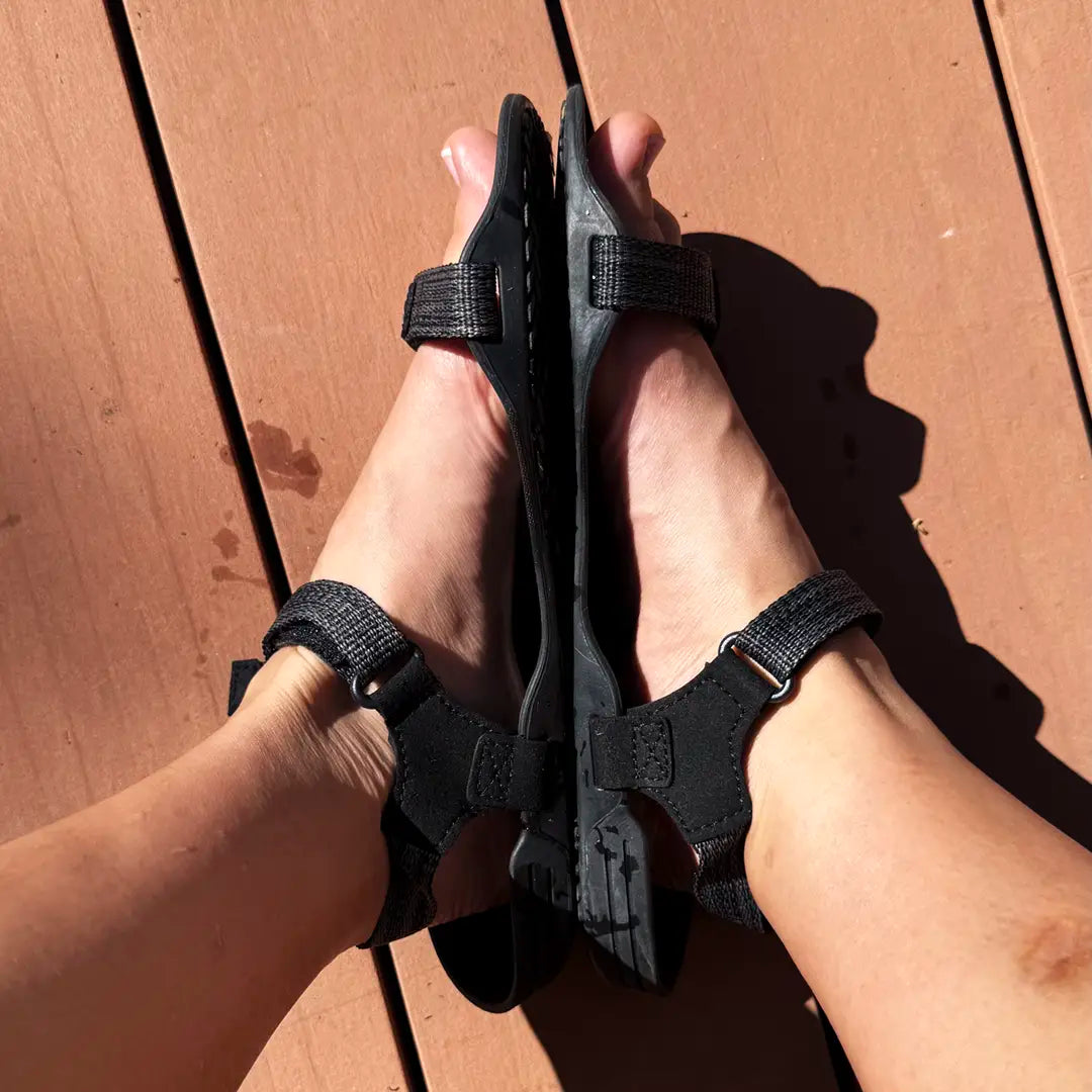 Persona usando sandalias Sowilo Raido negras sobre superficie de madera exterior.