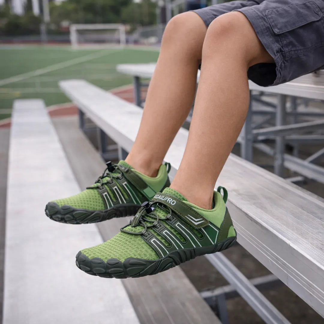 Niño sentado en el parque usando zapatillas barefoot verdes de malla transpirable.